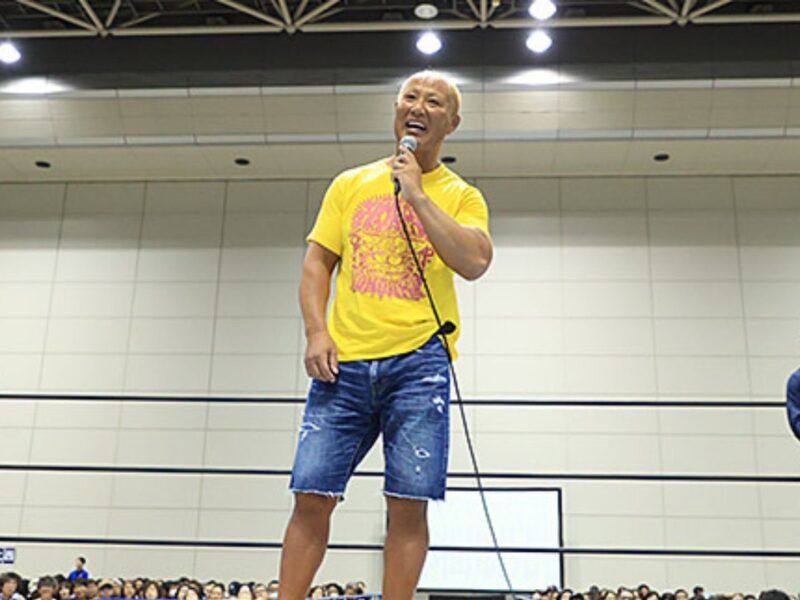 A man in a yellow T-shirt and blue shorts speaks into a microphone from the center of a boxing ring, with the audience visible around the ring.