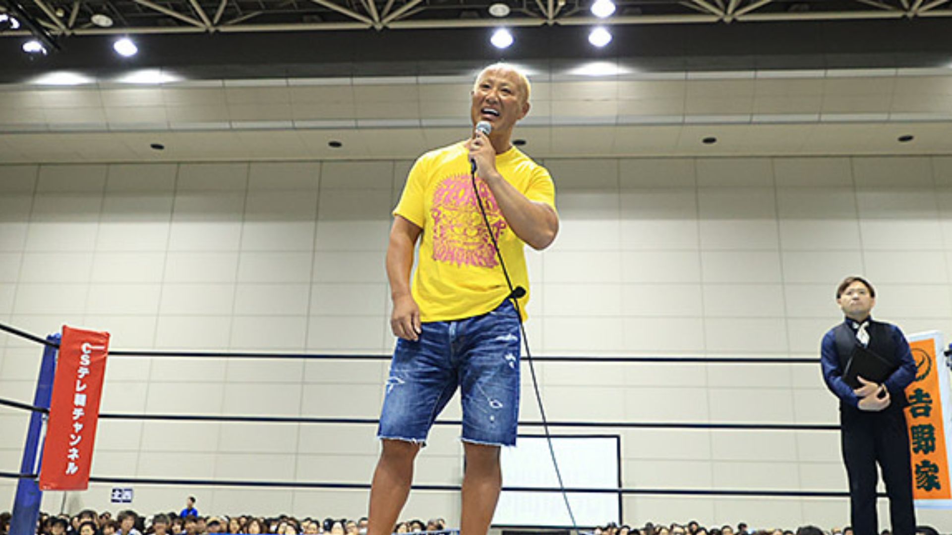A man in a yellow T-shirt and blue shorts speaks into a microphone from the center of a boxing ring, with the audience visible around the ring.