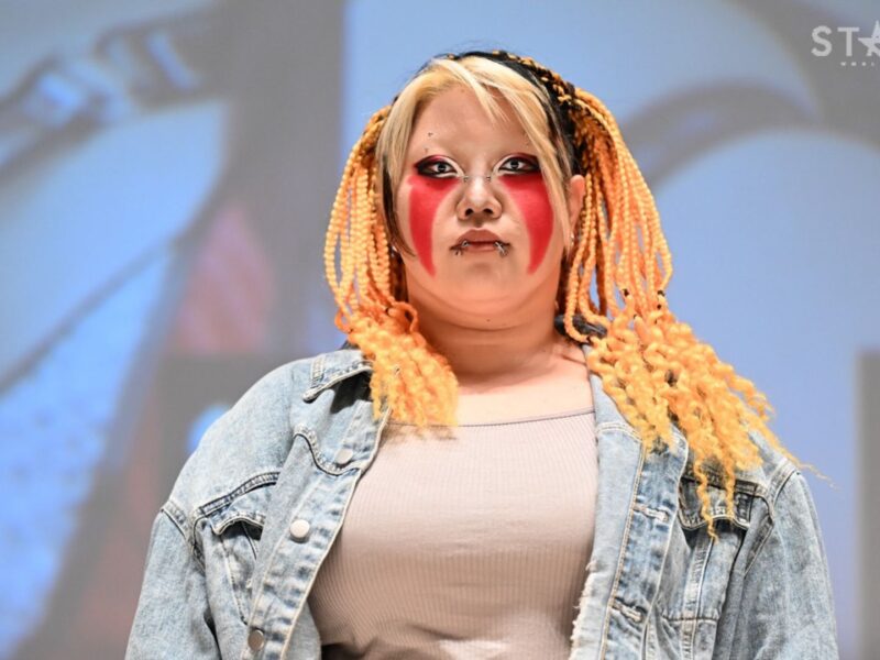 Model on stage with striking red facial makeup, orange braided hair, and a denim jacket on.