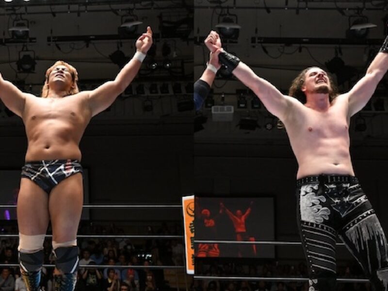 Two shirtless wrestlers in a ring raise their arms in victory as the crowd cheers in the background.