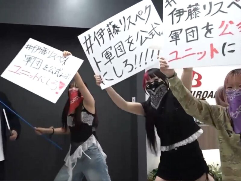 Group of protesters indoors, holding large signs with Japanese text; several wear masks or bandanas over their faces.