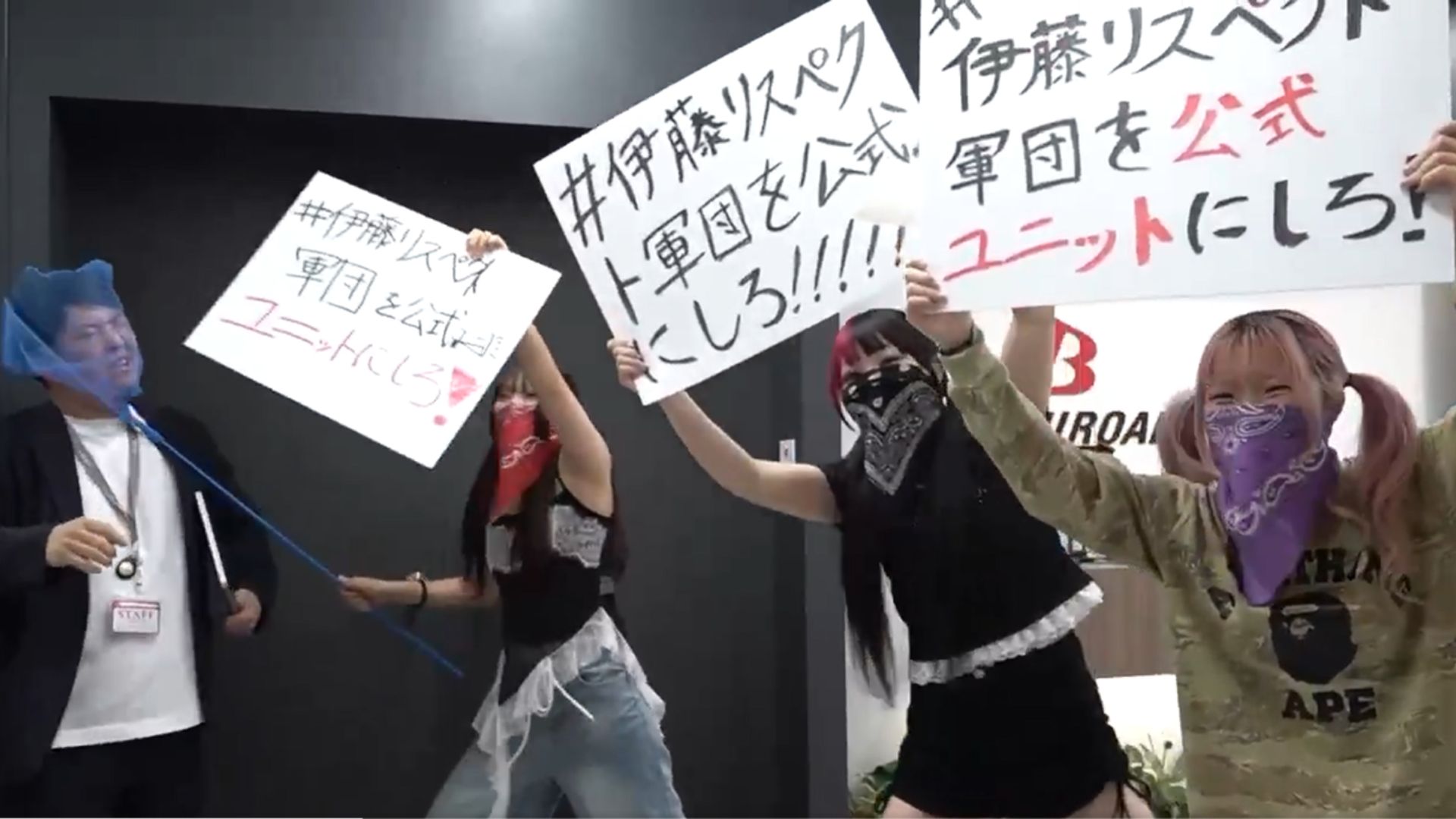 Group of protesters indoors, holding large signs with Japanese text; several wear masks or bandanas over their faces.
