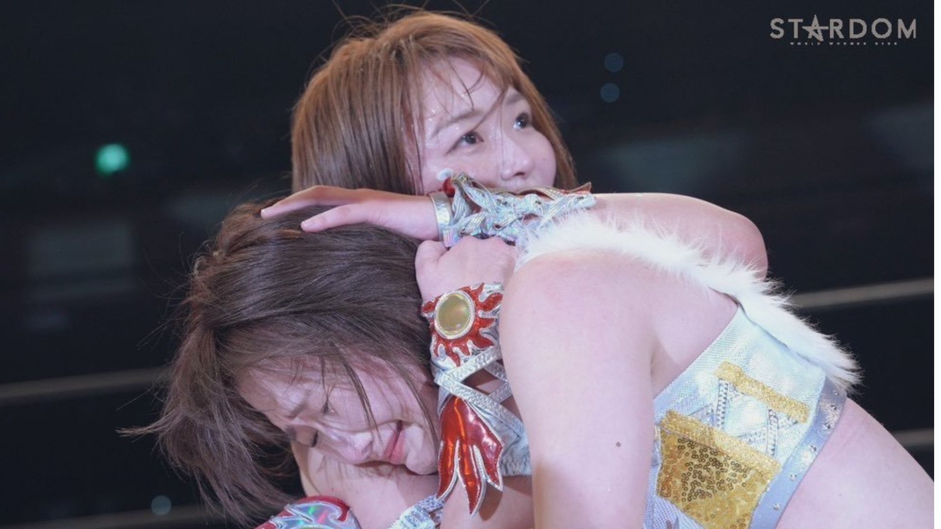 Two female performers share a hug on stage during a wrestling show; one face is visible, smiling, in a white and gold sequined outfit with accessories.