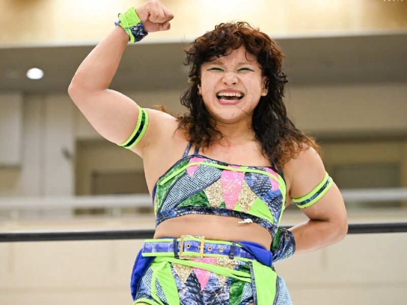 Female wrestler in neon-patterned outfit raises her fist in triumph inside a wrestling ring.