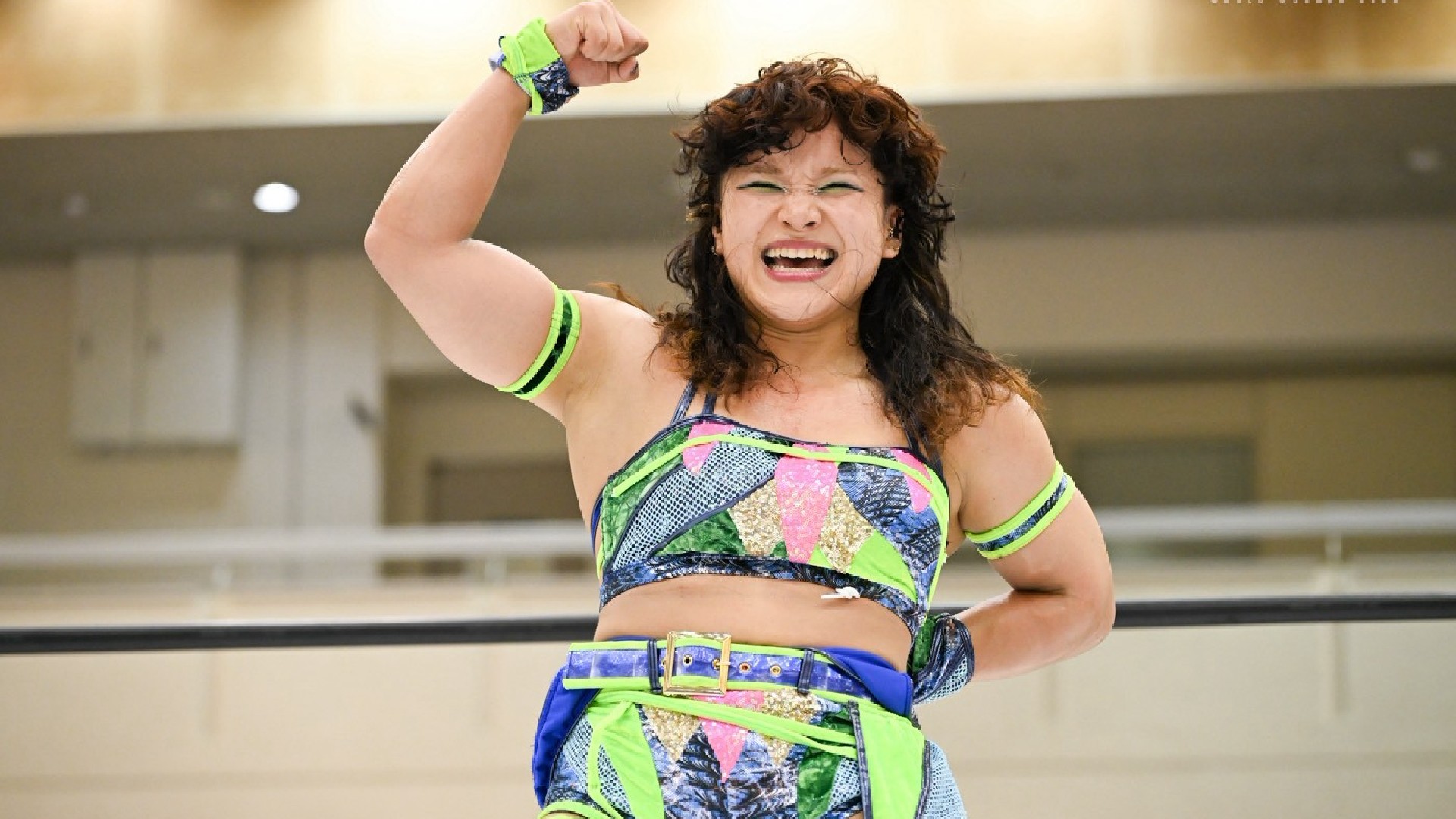 Female wrestler in neon-patterned outfit raises her fist in triumph inside a wrestling ring.
