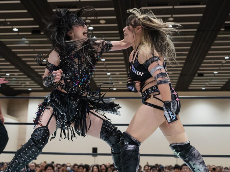 Two female wrestlers in black, embellished outfits clash in the ring as a referee watches from the left and a crowd looks on in the background.