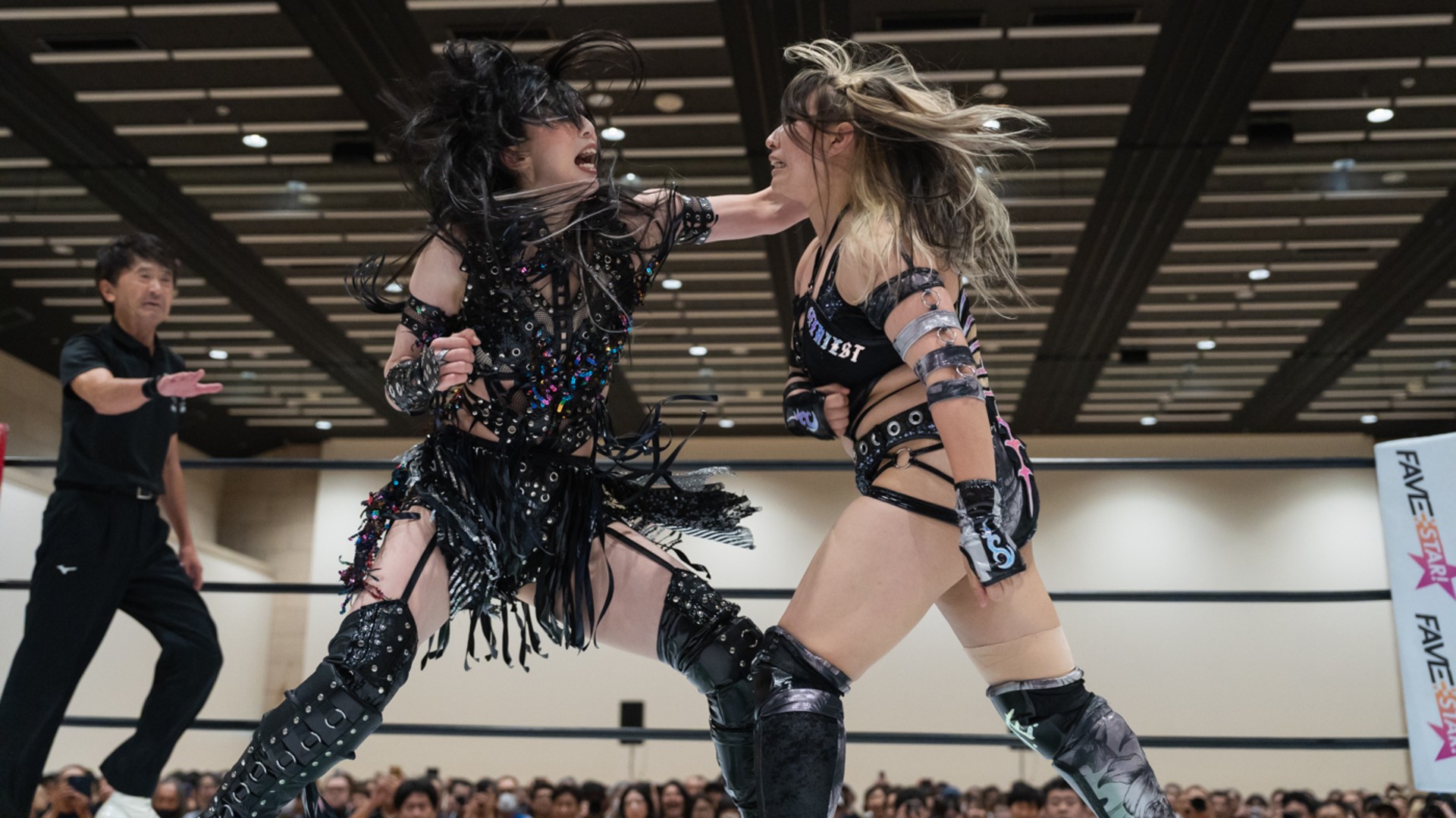 Two female wrestlers in black, embellished outfits clash in the ring as a referee watches from the left and a crowd looks on in the background.