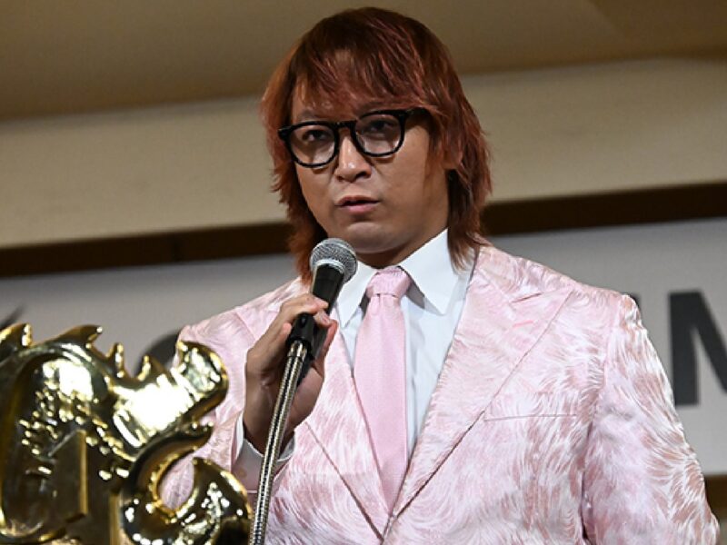 Man in a pink patterned suit and glasses speaks into a microphone on stage, with a large gold trophy in the foreground.