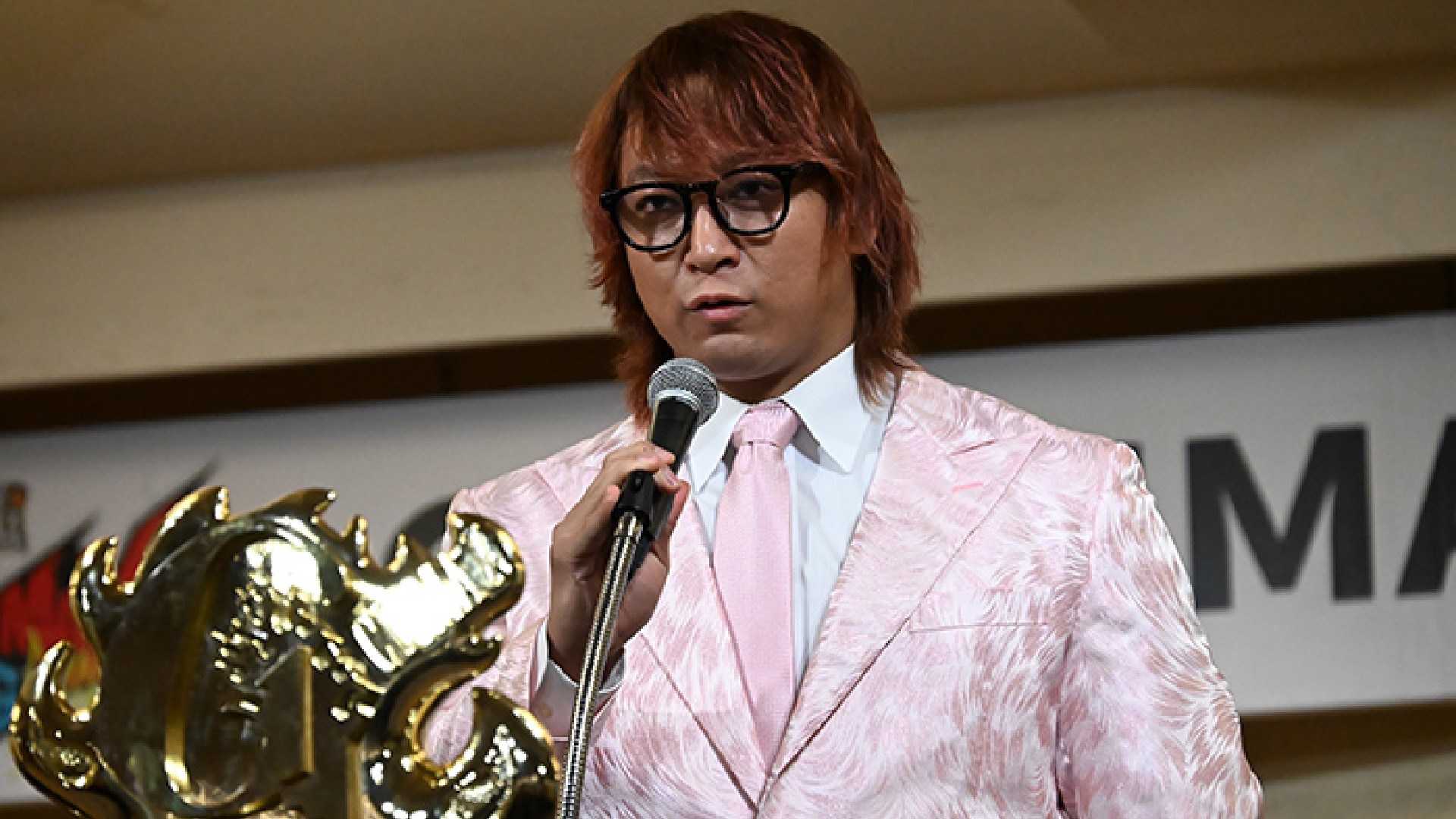Man in a pink patterned suit and glasses speaks into a microphone on stage, with a large gold trophy in the foreground.