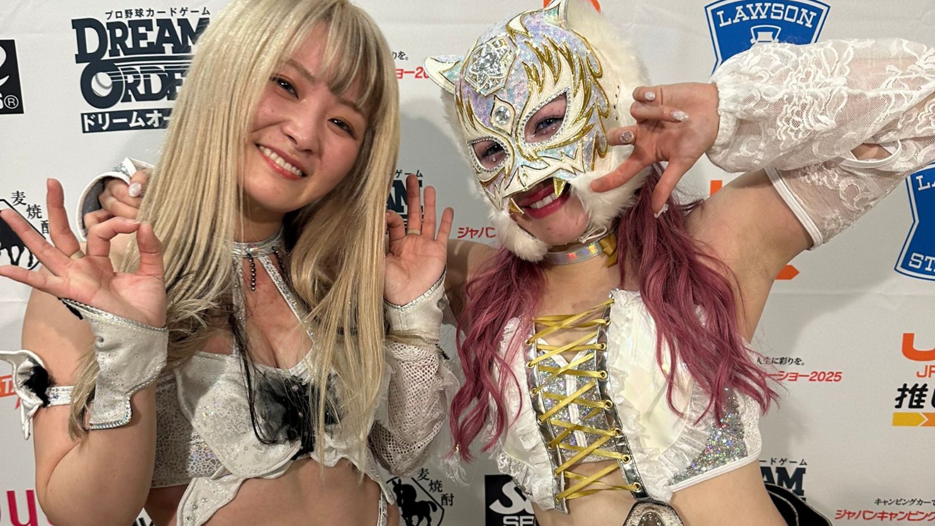 Two women in shiny costumes pose and smile at the camera, making peace signs in front of a sponsor backdrop.
