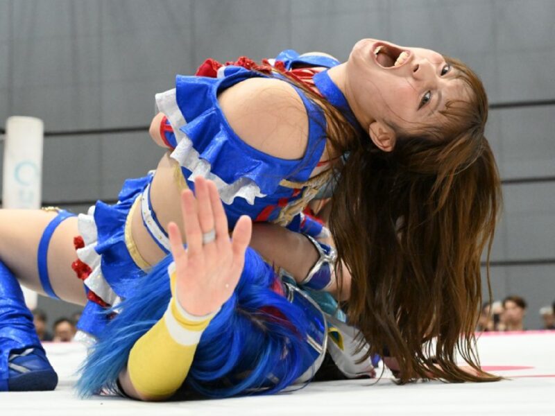Female wrestler in a blue outfit on a white mat during a match, long hair swinging as she leans back in an action move.