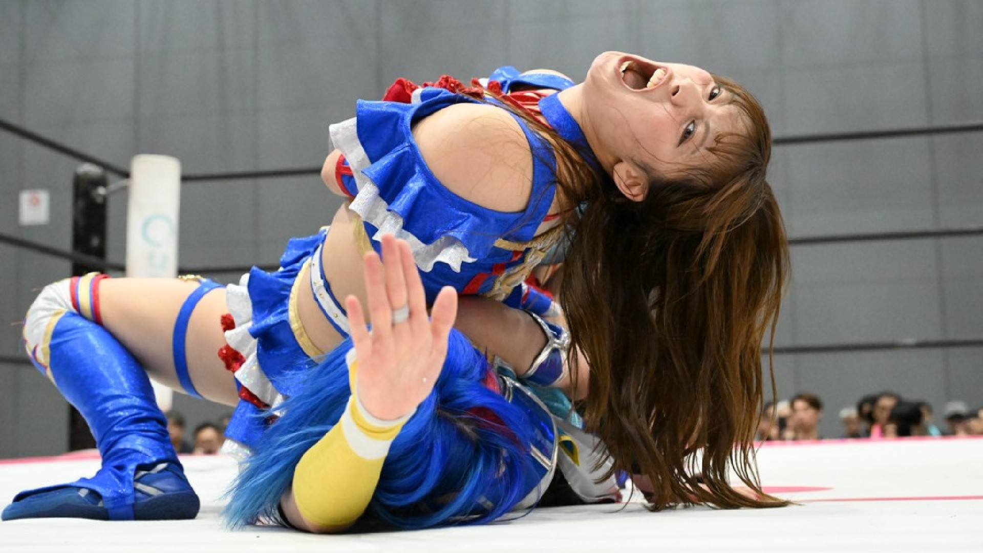 Female wrestler in a blue outfit on a white mat during a match, long hair swinging as she leans back in an action move.