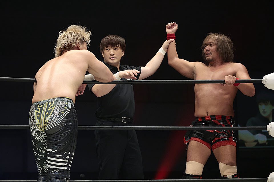 Referee raises the right wrestler's arm in victory as the left wrestler watches in the ring.