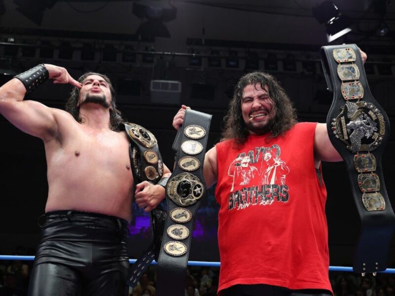 Two wrestlers celebrate in a ring with multiple championship belts displayed prominently.