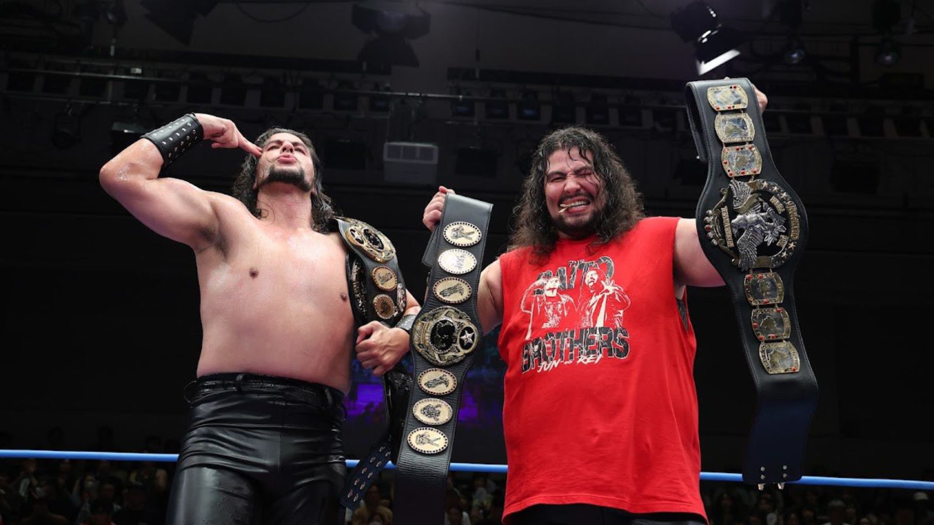 Two wrestlers celebrate in a ring with multiple championship belts displayed prominently.
