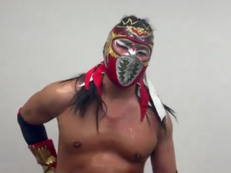 Lucha libre wrestler in a red and gold ornate mask, shirtless, standing with hands on hips against a plain wall.