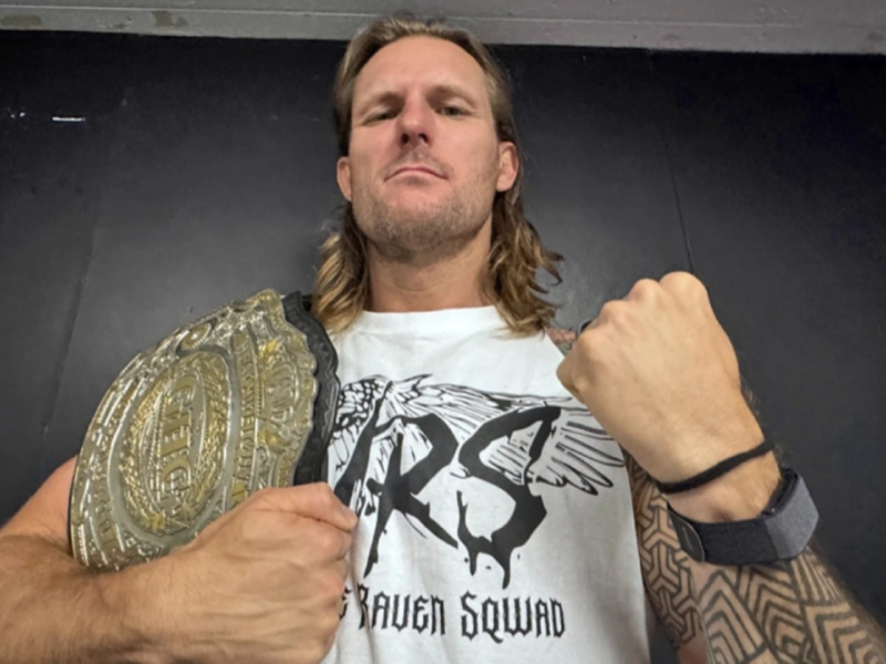 Wrestler with long blond hair holds a large championship belt over his shoulder, fist raised in a victory pose against a dark background, wearing a white graphic T‑shirt.