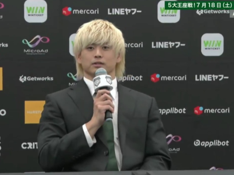 Man in a dark suit and green tie speaking into a microphone at a press conference with sponsor logos on a backdrop behind him.