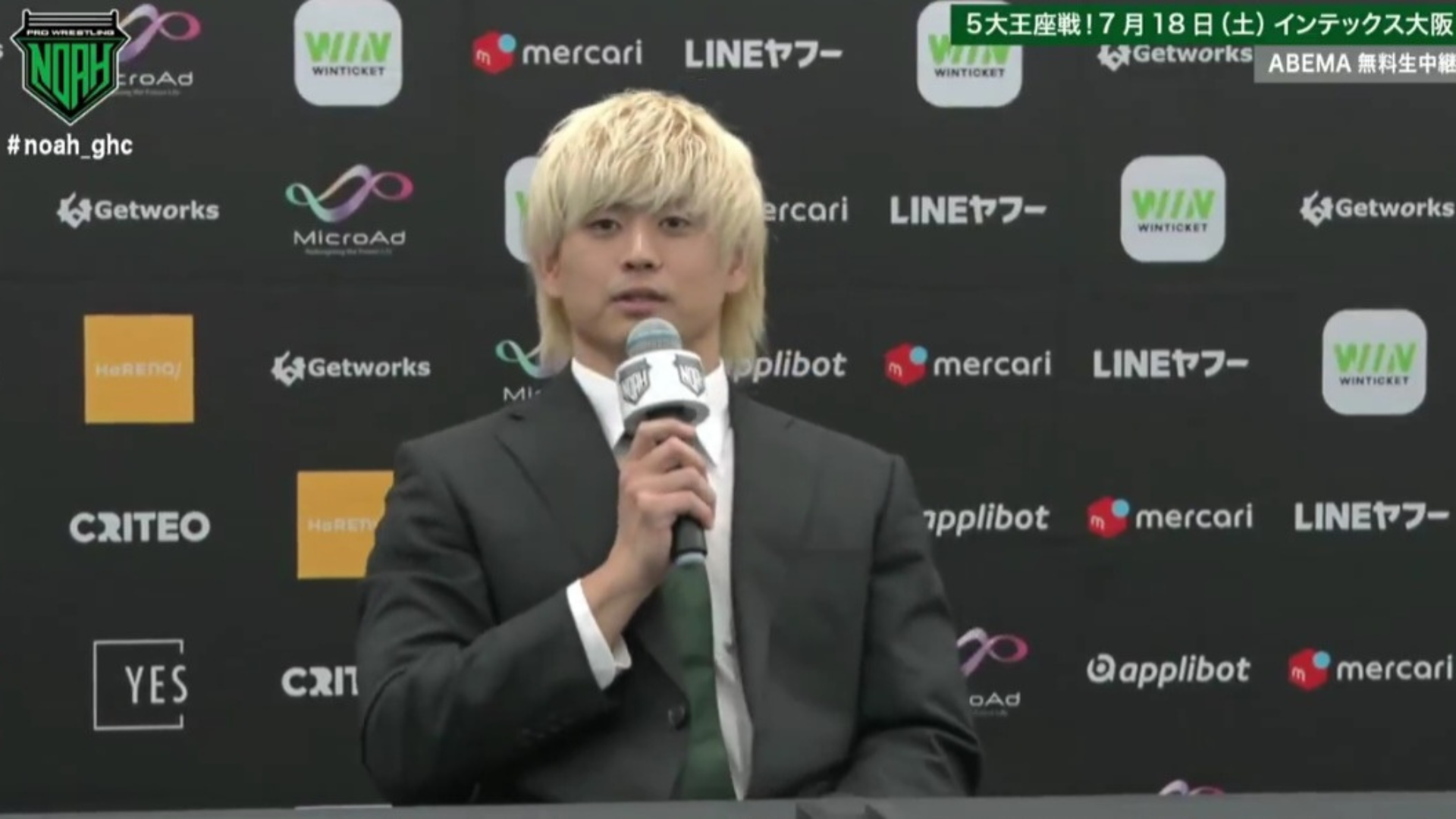 Man in a dark suit and green tie speaking into a microphone at a press conference with sponsor logos on a backdrop behind him.