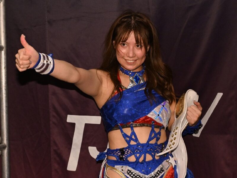 Female wrestler on stage giving a thumbs-up, holding a championship belt in a blue outfit.