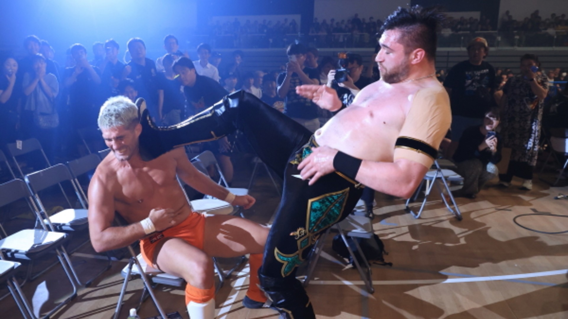 Wrestlers in a crowded gym: one shirtless man in orange shorts seated in a chair while another larger man in black and gold gear delivers a kick toward him. Audience and photographers watch from the background.