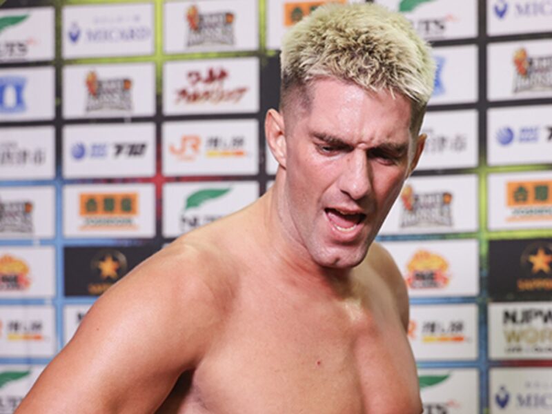 Shirtless man with bleached blond hair in front of a sponsor-filled backdrop at a press event.