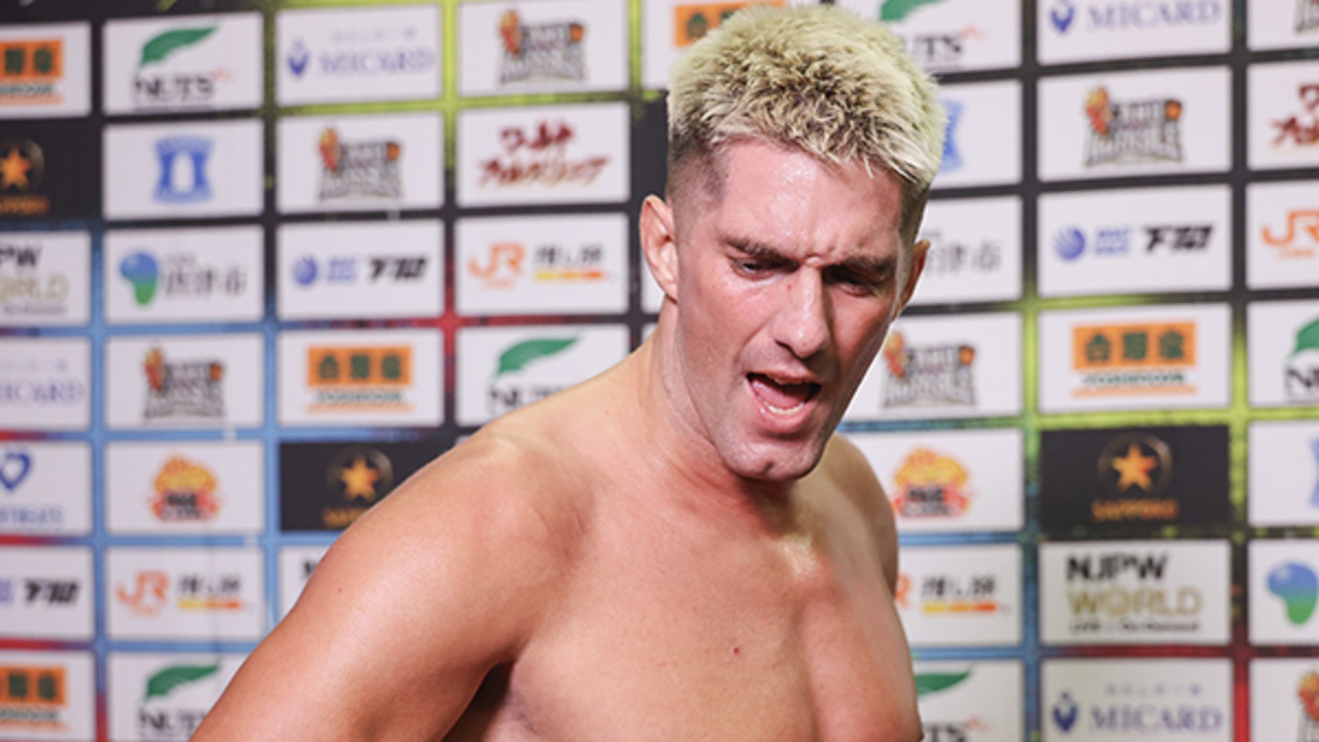 Shirtless man with bleached blond hair in front of a sponsor-filled backdrop at a press event.