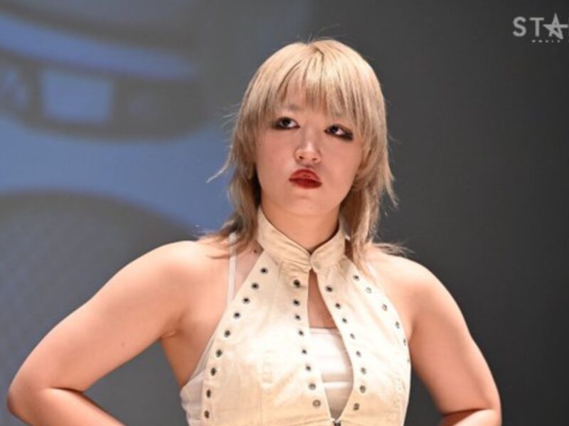 Female performer on stage with short blond hair, wearing a studded beige outfit and hands on hips, looking to the side (Stardom logo top right).