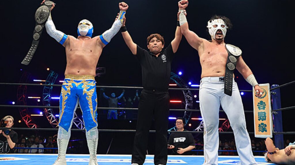 Two masked wrestlers stand with arms raised in victory as the referee holds them up inside a wrestling ring, belts draped over their shoulders.