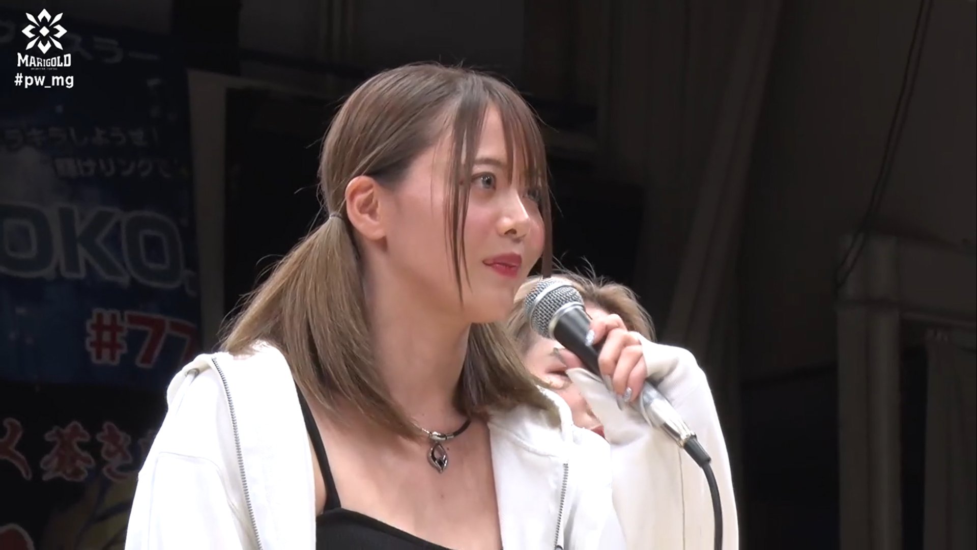 Woman with light brown hair holding a microphone and speaking on stage, wearing a black top and white jacket.