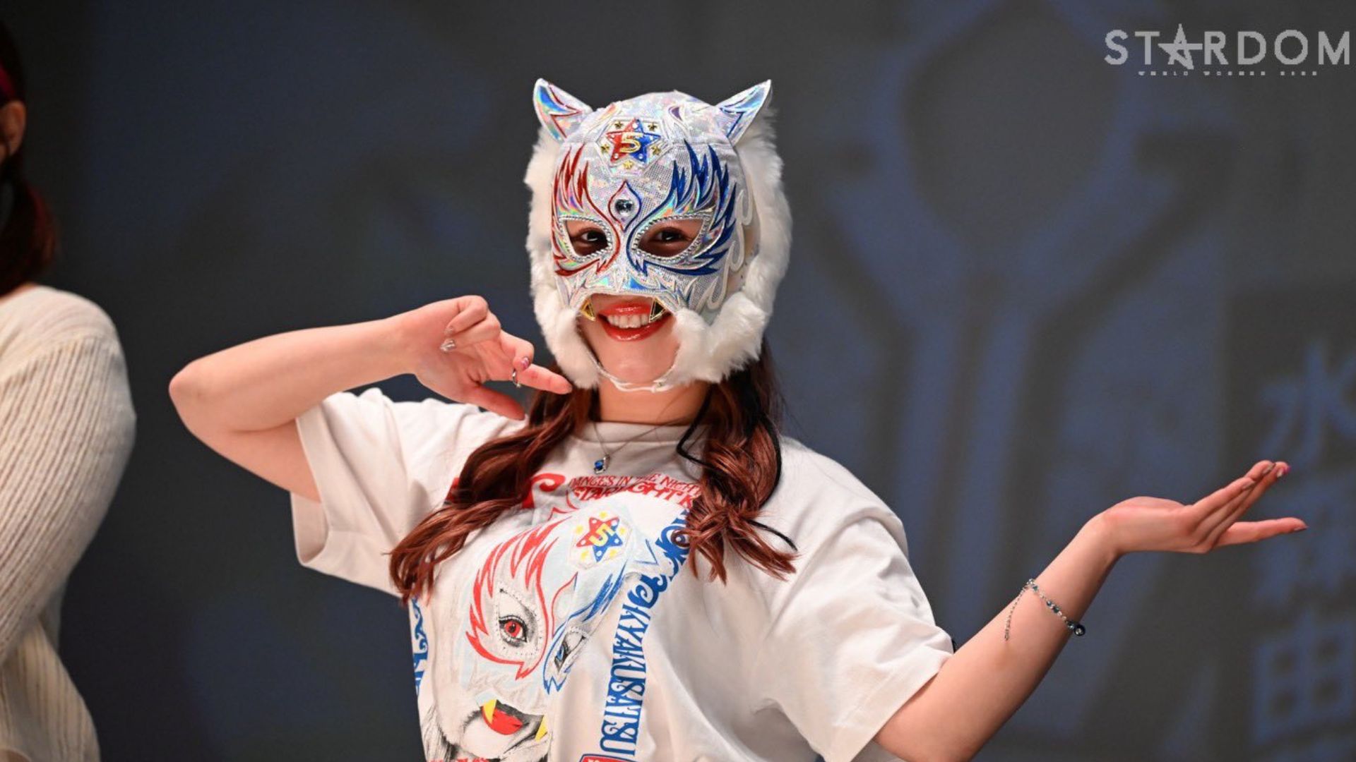 Person wearing a colorful cat mask with white fur, performing a pose on stage with hands raised