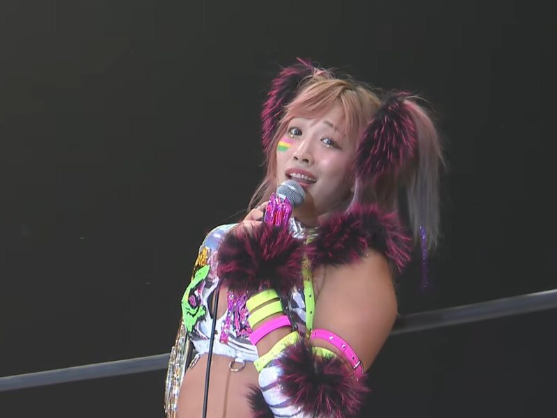 Performer in colorful outfit with neon accessories singing into a microphone on stage, with a dark background and logo in the corner.