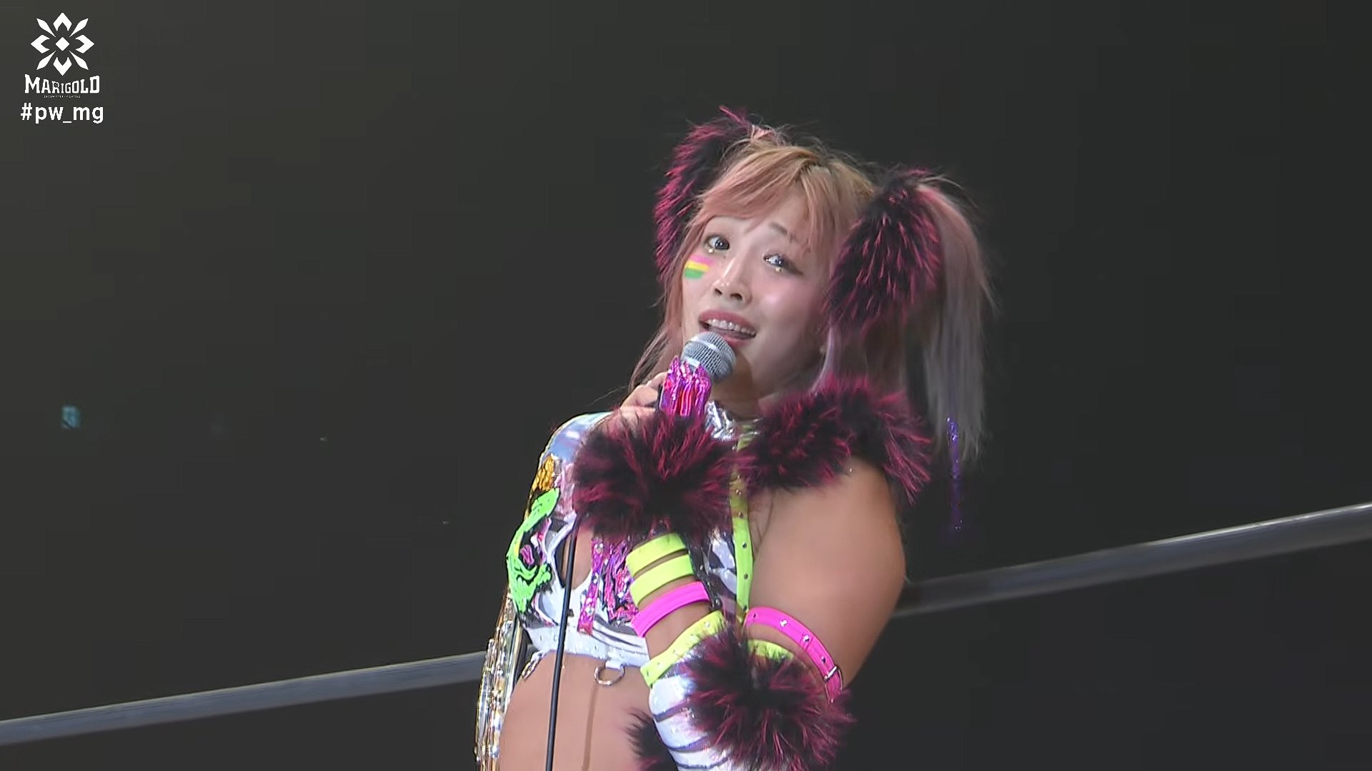 Performer in colorful outfit with neon accessories singing into a microphone on stage, with a dark background and logo in the corner.