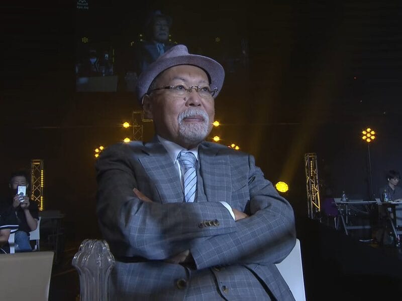 Older man in a gray plaid suit and hat sits with arms crossed on a stage, stage lights behind him.