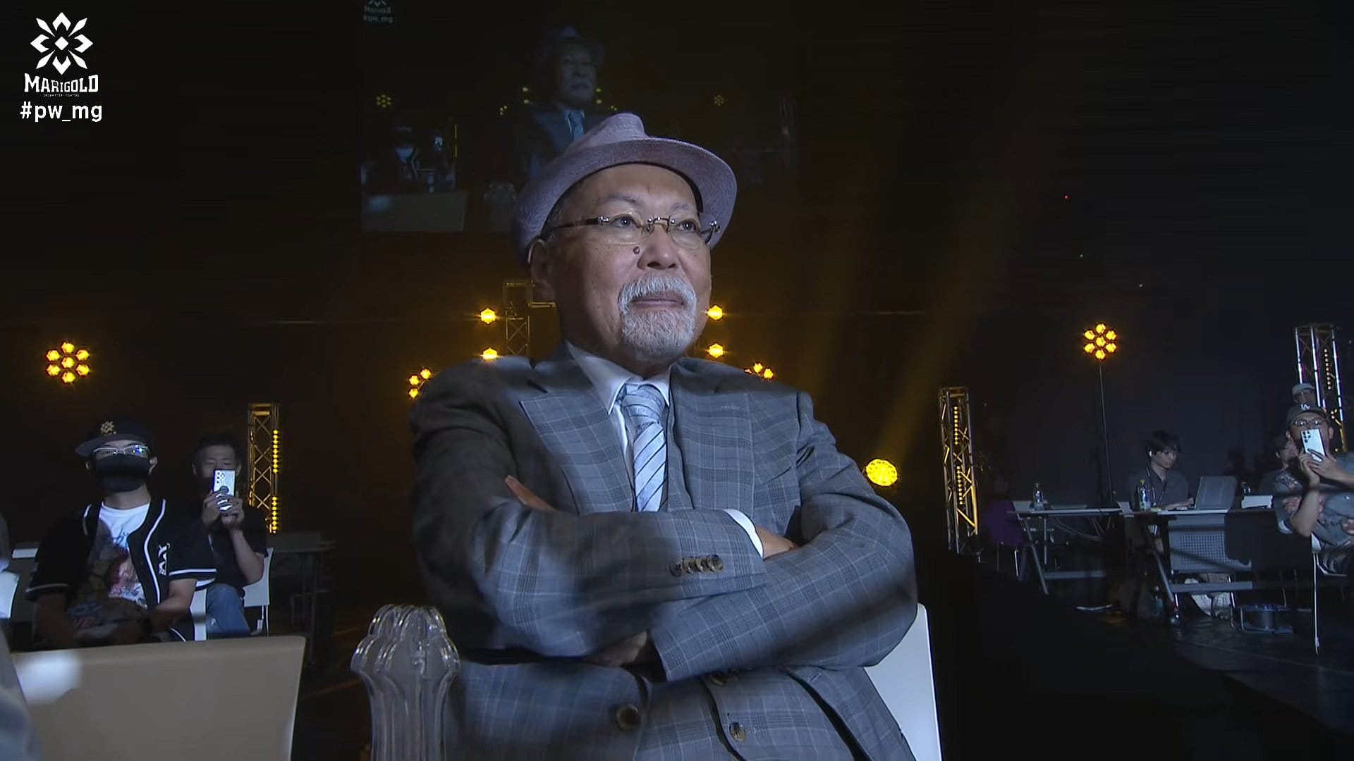 Older man in a gray plaid suit and hat sits with arms crossed on a stage, stage lights behind him.