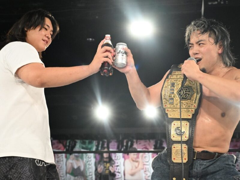 Two wrestlers in a ring toast with bottles, one wearing a gold championship belt and microphone.