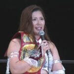 Female professional wrestler in red and white outfit holding a microphone, with a gold championship belt draped over her shoulder on stage against a dark background.