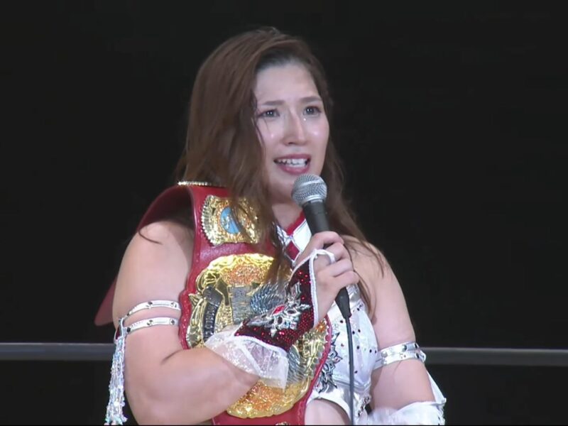 Female professional wrestler in red and white outfit holding a microphone, with a gold championship belt draped over her shoulder on stage against a dark background.