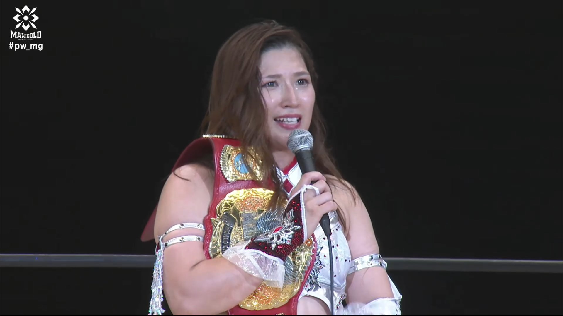 Female professional wrestler in red and white outfit holding a microphone, with a gold championship belt draped over her shoulder on stage against a dark background.