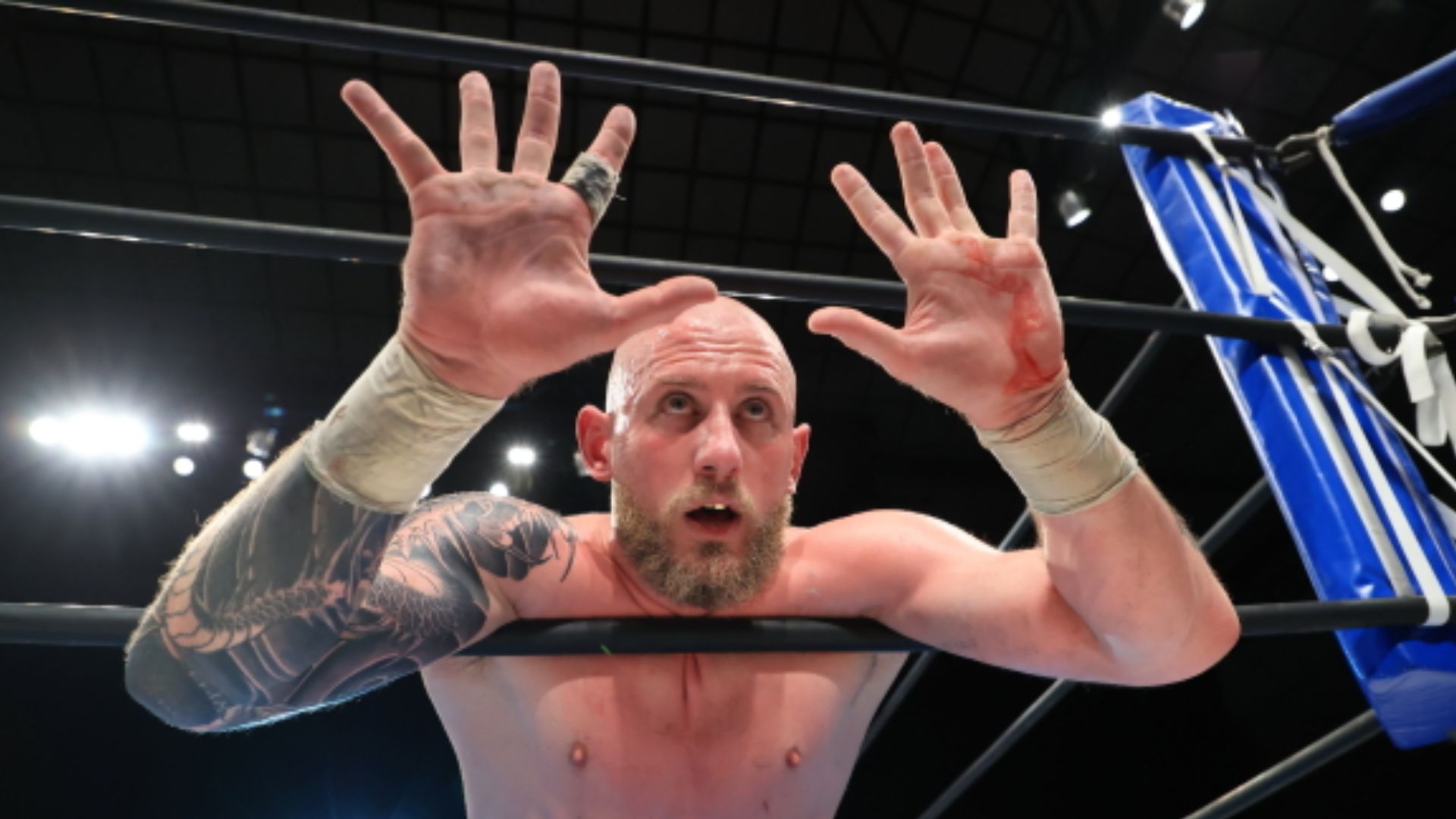 Wrestler with a tattooed arm leans over the ropes, hands raised to the camera, a cut showing red on his right palm.