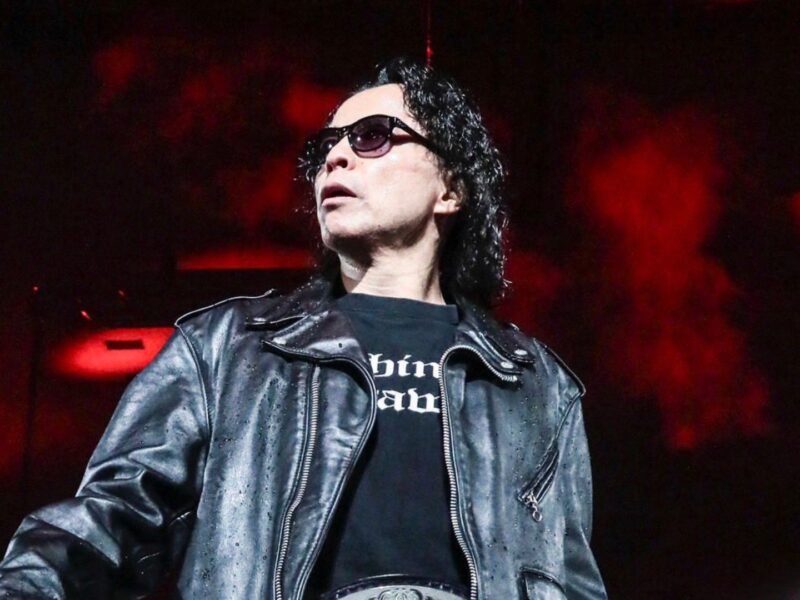 Male singer in a black leather jacket and sunglasses performing on a dark stage with red lights.