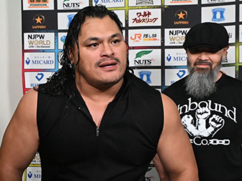 Two muscular men stand side by side in front of a sponsor backdrop; the left man has long dark hair and a sleeveless black shirt, the right man wears a cap and has a gray beard.