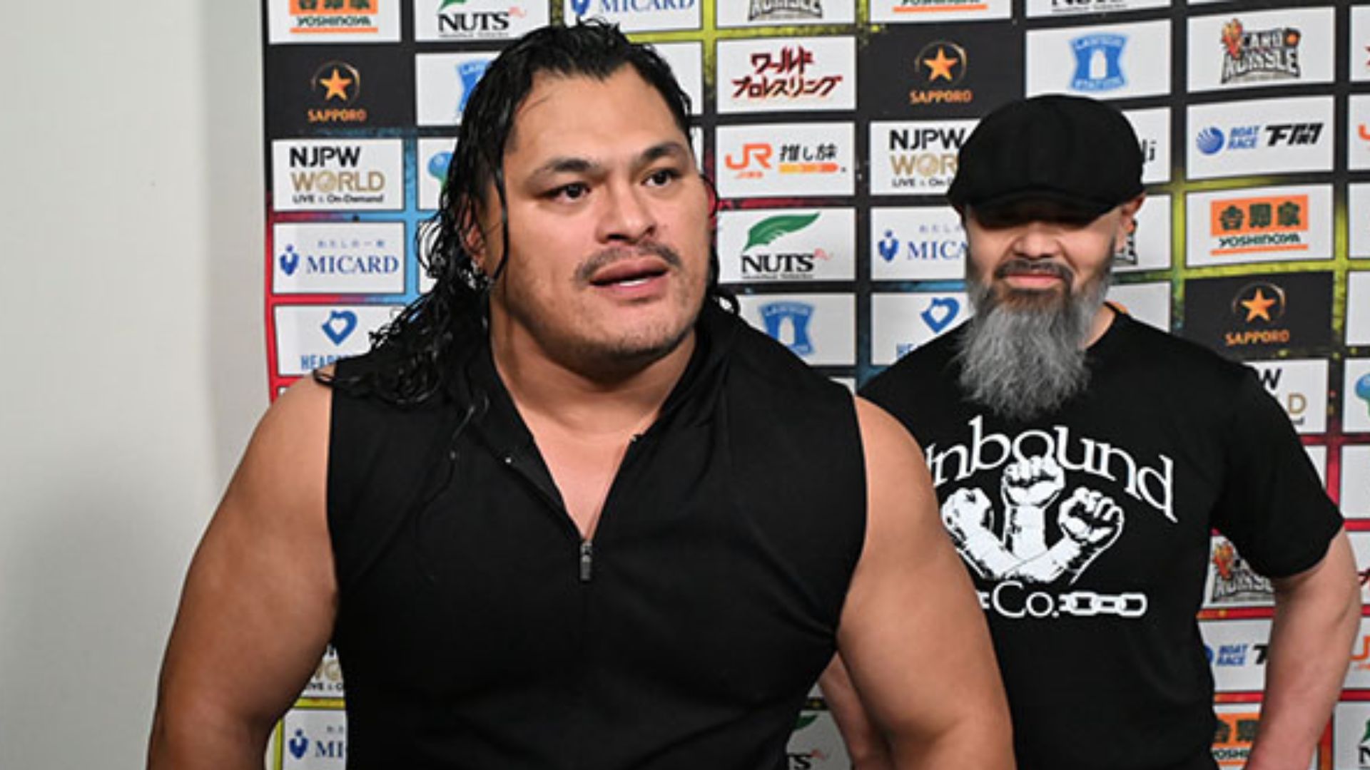 Two muscular men stand side by side in front of a sponsor backdrop; the left man has long dark hair and a sleeveless black shirt, the right man wears a cap and has a gray beard.