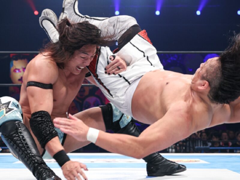 Two male wrestlers collide mid-air in a ring as one executes a flying kick toward the other, with a referee and audience in the background.