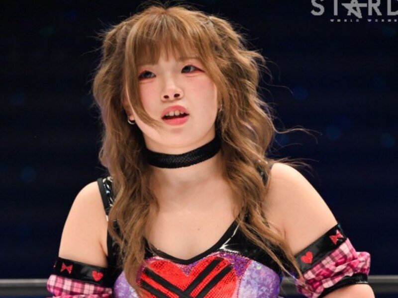 Female professional wrestler with light brown hair, wearing a black choker and colorful red/black outfit with plaid sleeves, in a wrestling ring during a Stardom event.