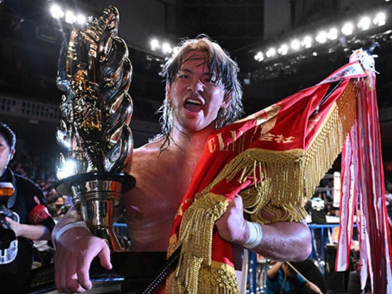 Wrestler holding a tall trophy, wearing a red and gold cape, cheering in a crowded arena with photographers nearby