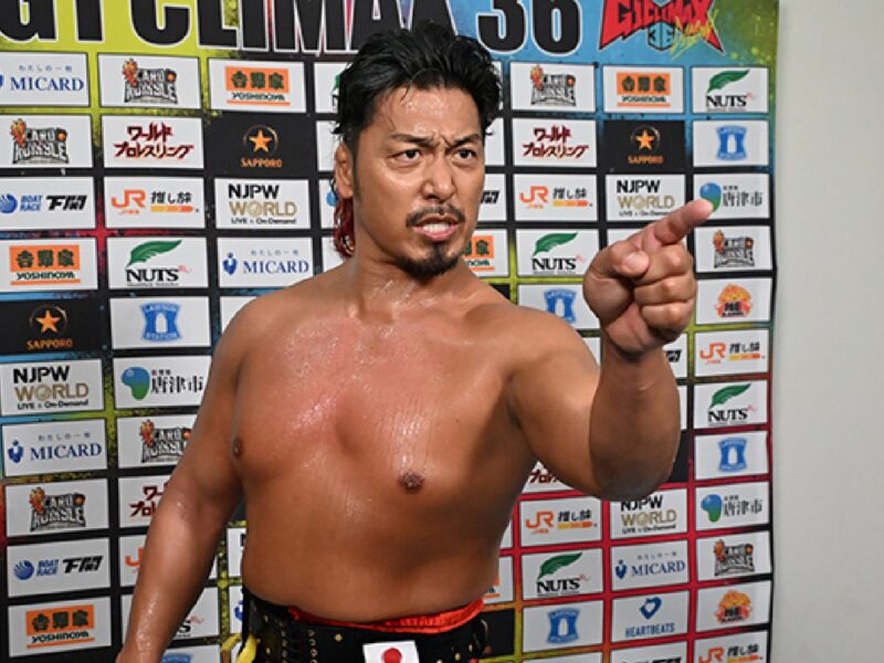 Shirtless male wrestler with wet hair points at the camera in a sponsor-filled background.