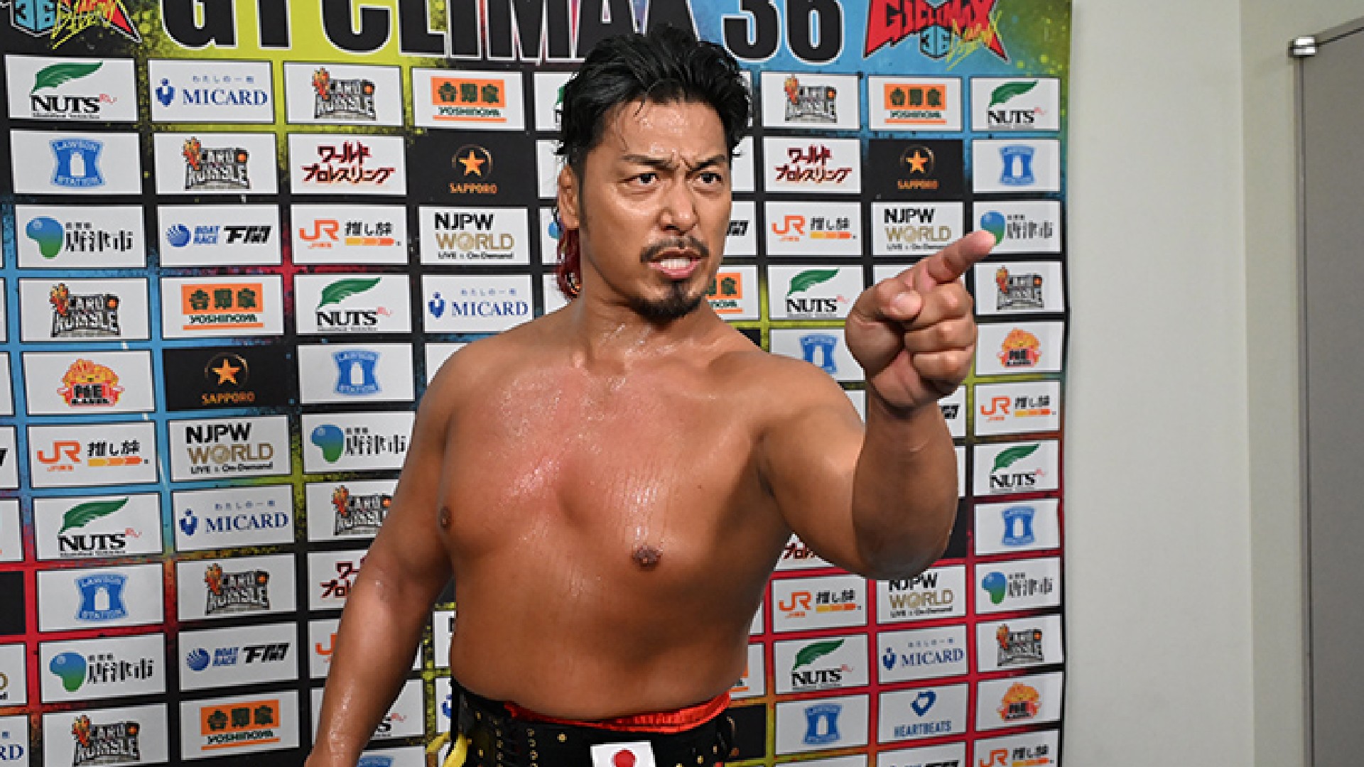 Shirtless male wrestler with wet hair points at the camera in a sponsor-filled background.
