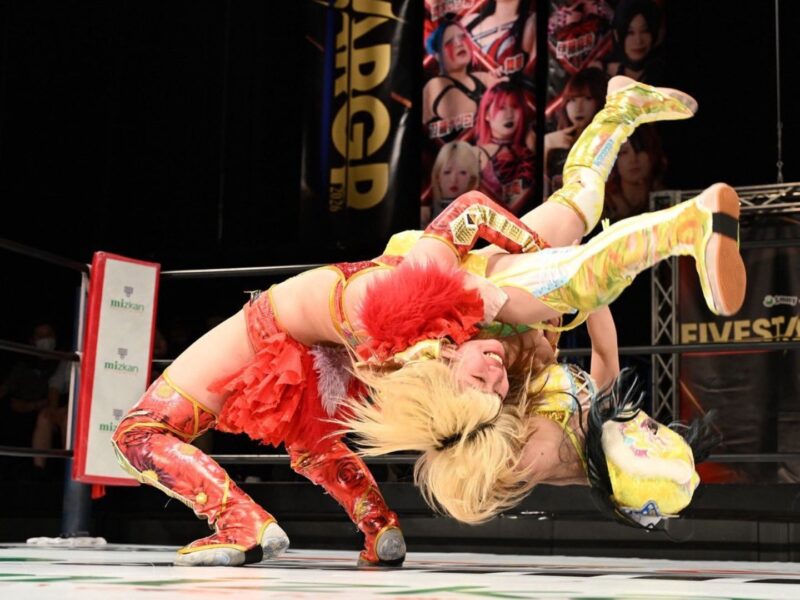 Two female wrestlers in bright red and yellow outfits collide mid-air in a wrestling ring, crowd and banners visible in the background