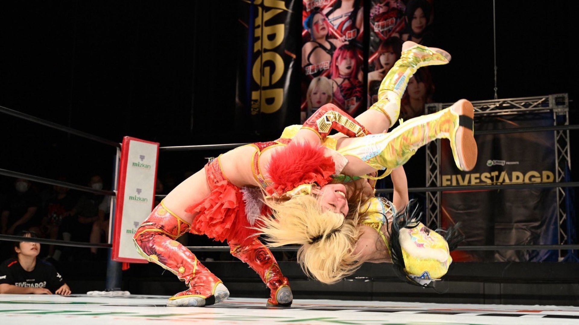 Two female wrestlers in bright red and yellow outfits collide mid-air in a wrestling ring, crowd and banners visible in the background